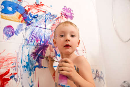 Portrait of cute adorable white Caucasian little boy playing and painting with paints on wall in bathroom having fun lifestyle childhood conceptの写真素材