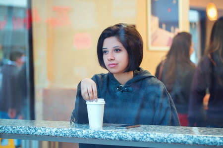 Candid portrait of beautiful young hipster latin hispanic girl woman with short hair bob in grey cape poncho jacket sitting in cafe restaurant with coffee looking away shot through windowの写真素材