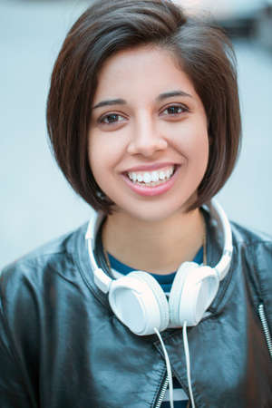 Portrait of beautiful Hispanic latin girl woman short black hair in leather jacket with headphones, outside in evening night city street, smiling laughing looking in camera, lifestyle portrait conceptの写真素材