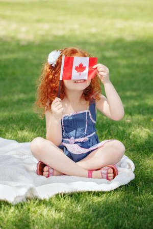 Portrait of  cute little red-haired Caucasian girl child holding Canadian flag with red maple leaf, sitting on grass in park outside, celebrating Canada Day anniversaryの写真素材