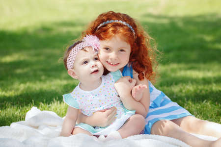 Portrait of two cute adorable little red-haired Caucasian girls sisters siblings children, sitting together in field meadow park outside, hugging, looking in camera, happy lifestyle childhood conceptの写真素材