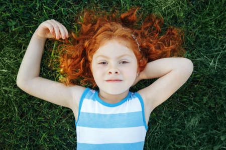 Portrait of cute adorable pensive little red-haired Caucasian girl child in blue dress lying  on green grass in park outside dreaming thinking, happy lifestyle childhood concept, view from aboveの写真素材