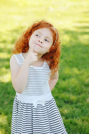 Portrait of cute adorable puzzled little red-haired Caucasian girl child in white striped dress standing in field meadow park outside thinking, having fun, happy lifestyle childhood conceptの写真素材