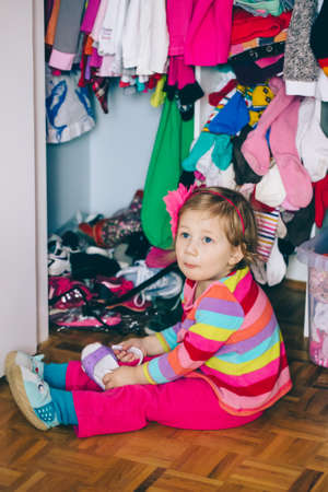 Portrait of cute adorable  little white Caucasian girl baby sitting on floor in wardrobe, wearing shoes, funny emotional face expression, lifestyle authentic childhood conceptの写真素材