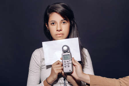 Portrait of beautiful hispanic latin brunette young woman in studio holding white paper and flash meter, measuring light, on plain black backgroundの写真素材