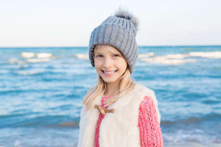 Portrait of smiling blonde white Caucasian child kid girl with long hair wearing white fur jacket gilet and grey hat, on sea shore beach looking in camera, happy lifestyle childhoodの写真素材