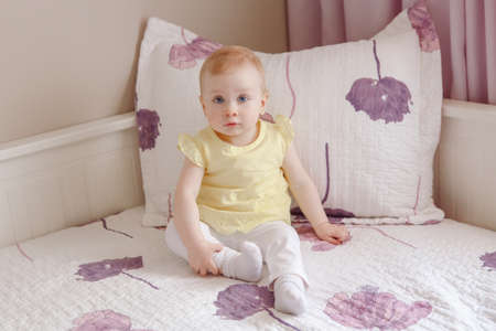 Portrait of cute adorable white blonde Caucasian smiling baby girl with large blue eyes in yellow shirt, sitting on bed, looking in cameraの写真素材
