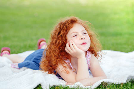 Portrait of cute funny happy smiling little red-haired Caucasian girl child in blue dress lying on green grass in park outside, dreaming thinking, lifestyle childhood conceptの写真素材