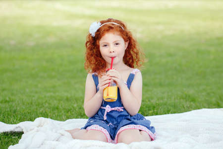 Portrait of cute adorable little red-haired Caucasian girl child in blue dress sitting on white blanket in park outside drinking orange juice from glass jar, happy lifestyle childhood conceptの写真素材