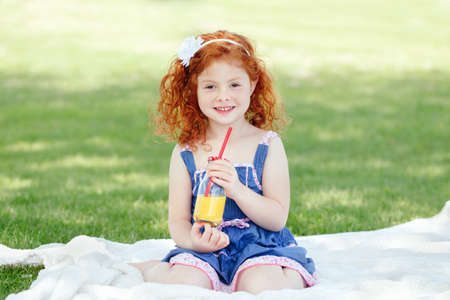 Portrait of cute adorable smiling little red-haired Caucasian girl child in blue dress sitting on white blanket in park outside with glass jar of orange juice, happy lifestyle childhood conceptの写真素材