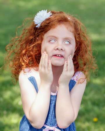 Portrait of cute adorable little red-haired Caucasian girl child in blue dress making funny scary silly faces in park outside, child rolling eyes, having fun, lifestyle childhood conceptの写真素材