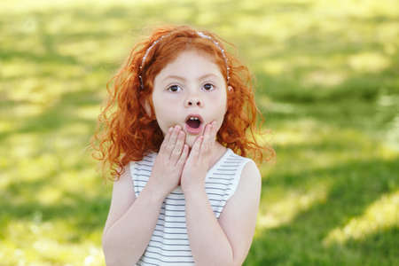 Portrait of cute adorable surprised  little red-haired Caucasian girl child in striped dress in park outside, playing  crying screaming in fear, happy lifestyle childhood conceptの写真素材