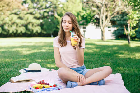 Portrait of young beautiful white Caucasian woman girl sitting on grass in park with closed eyes drinking fruit juice on summer day, toned with retro filters, film effect, rural country lifestyleの写真素材