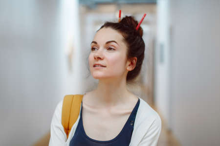 Portrait of smiling laughing white Caucasian brunette young beautiful woman student with pencils in hair bun, standing in hall of college university, artist designer at workの写真素材