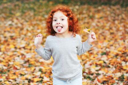 Portrait of cute adorable little red-haired Caucasian girl child making funny silly faces, showing tongue, in autumn fall park outside, playing having fun, lifestyle childhoodの写真素材