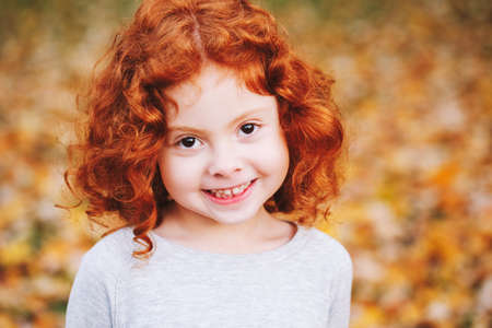 Closeup portrait of cute adorable smiling  little red-haired Caucasian girl child standing in autumn fall park outside, looking in camera, happy lifestyle childhood conceptの写真素材