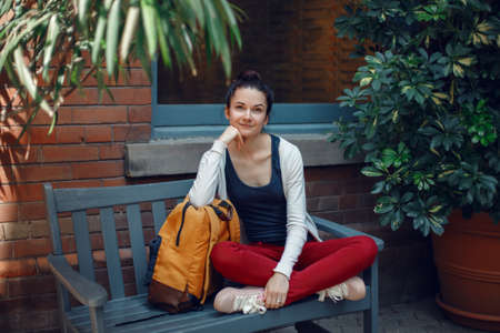 Portrait of smiling beautiful Caucasian young girl woman in white sweater and red jeans, sitting with yellow travel bag backpack on bench in park, looking in camera, wanderlust adventure vacationの写真素材