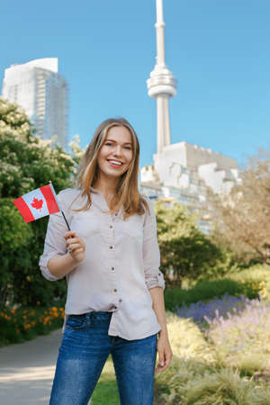 Portrait of beautiful smiling white blonde Caucasian woman holding waving Canadian flag with red maple leaf, outside in Toronto city near CN Tower, looking in camera, celebrating Canada Dayの写真素材