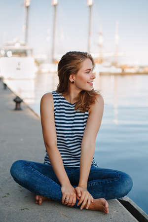 Portrait of white Caucasian blonde woman with tanned skin striped t-shirt and blue jeans sitting on pier by seashore lakeshore, with yacht boat ship on background on water, lifestyle summerの写真素材