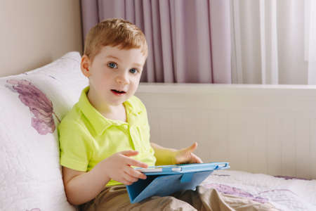 Portrait of cute adorable white Caucasian toddler boy sitting in bed playing with digital tablet looking in camera with funny face expression. Lifestyle early development. New technology generation.の写真素材