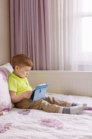 Portrait of cute adorable white Caucasian toddler boy sitting in bed playing with digital tablet with funny face expression. Candid lifestyle early development. New technology generation.の写真素材