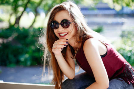 Portrait of beautiful sexy smiling white Caucasian girl woman with long blond red hair, wearing jeans and t-shirt, sitting on bench in park outside with sunglasses. Hot summer day, lifestyle concept.の写真素材
