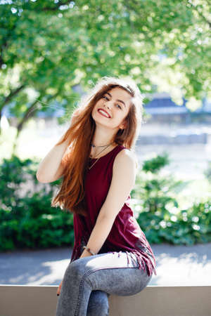 Portrait of beautiful sexy smiling white Caucasian girl woman with long blond red hair, wearing jeans and t-shirt, sitting on bench in park outside with sunglasses. Hot summer day, lifestyle concept.の写真素材