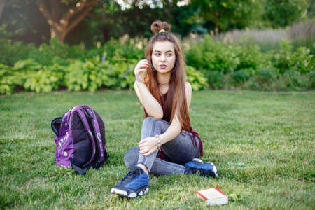 Portrait of beautiful pensive white Caucasian young woman student female, sitting on grass  in park outside with backpack and book. Hobby education back-to-school concept.の写真素材
