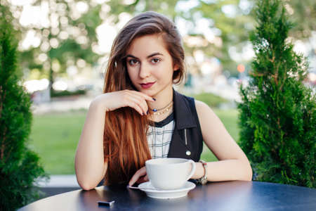 Portrait of beautiful smiling young white middle eastern Caucasian girl woman with long hair sitting at table, looking in camera. Woman with cup of coffee tea outside on summer day. の写真素材