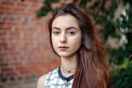 Closeup portrait of beautiful pensive young middle eastern Caucasian jewish woman with long dark hair, black brown eyes, looking in camera. Girl in white plaid shirt against brick wall. Natural beautyの写真素材