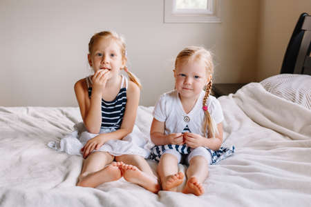 Portrait of two cute adorable little red-haired blonde Caucasian girls sisters sitting together on bed at home. Siblings looking in camera. Happy lifestyle childhood concept.の写真素材