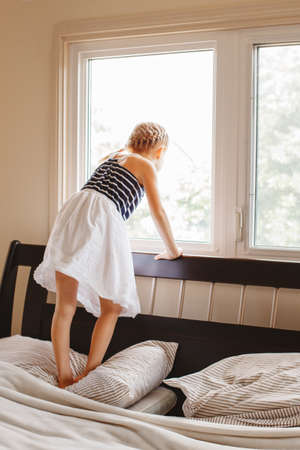 Portrait of white Caucasian child girl standing on bed at home looking out of window outside. View from back.の写真素材