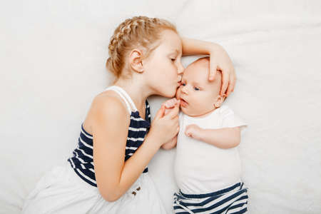 Lifestyle portrait of cute white Caucasian girl sister holding kissing little baby, lying on bed indoors. Older sibling with younger brother newborn. Family love bonding together concept.の写真素材