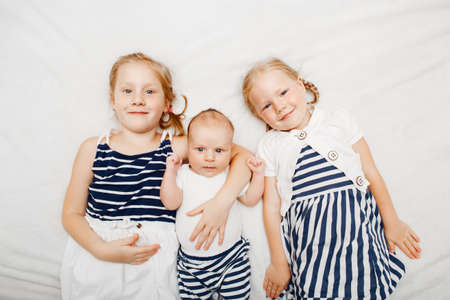 Lifestyle portrait of cute white Caucasian girls sisters holding little baby, lying on bed blanket indoors. Older siblings with younger brother sister newborn. Family love bonding together concept.の写真素材