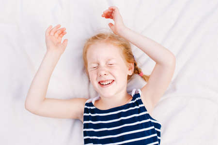 Closeup lifestyle portrait of white Caucasian red-haired girl smiling laughing. Kid lying with closed eyes on white blanket. View from top above. Happy childhood dreams, enjoying life concept.の写真素材