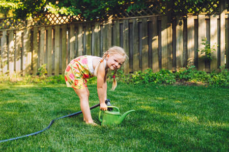 Portrait of little girl filling watering pot from gardening house on backyard on summer day. Child with watering can in garden. Lifestyle family activity. Kids responsibility for doing home chores.の写真素材