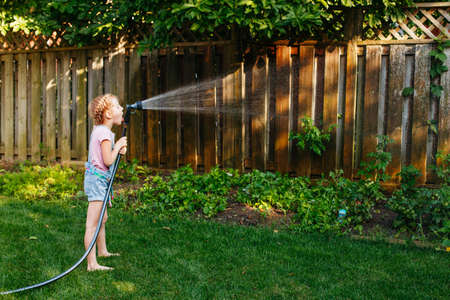 Portrait of girl watering plants vegetables with gardening house on backyard on summer day. Child playing drinking  water outside. Lifestyle family activity. Kids responsibility for doing home chores.の写真素材