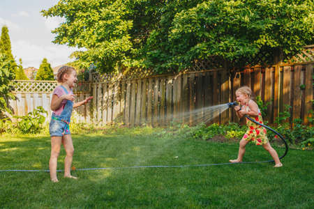 Two girls splashing each other with gardening house on backyard on summer day. Children playing with water outside at sunset. Candid moment, lifestyle activity. Life friendship of sisters siblings.の写真素材