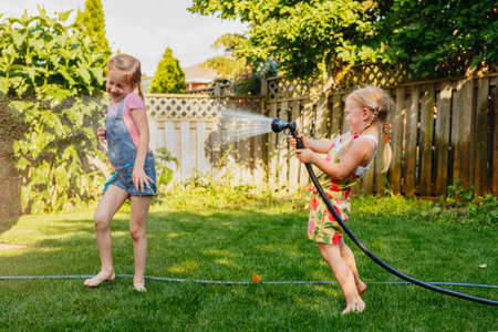 Two girls splashing each other with gardening house on backyard on summer day. Children playing with water outside at sunset. Candid moment, lifestyle activity. Life friendship of sisters siblings.の写真素材