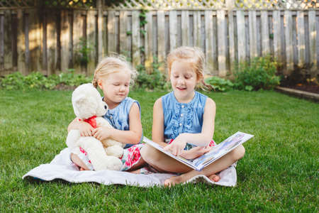 Group portrait of of two cute white Caucasian children friends sitting on grass outside with book. Preschool girls sisters reading book on backyard. Back to school fall concept.の写真素材
