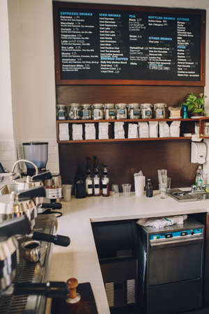 Interior of coffee shop. Wooden board with names of coffee hot and cold drinks, prices. Glass jars with tea bags on shelves. Professional espresso coffee maker. Toned with retro vintage filters.の写真素材