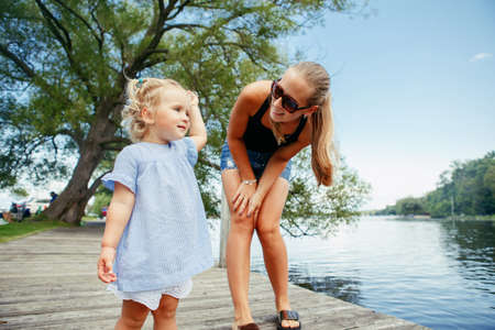 Group portrait of happy white Caucasian mother and daughter child having fun outside. Mom and child girl playing, hugging in park on pier by water lake. Candid authentic real lifestyle.の写真素材