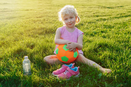 Portrait of happy cute adorable white Caucasian girl holding soft soccer ball. Child sitting in grass outside after playing soccer football game on summer day. Healthy lifestyle childhood concept.の写真素材
