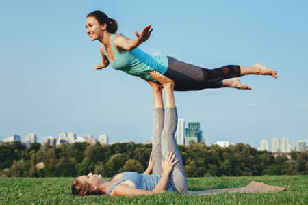Two young beautiful Caucasian women yogi doing acro bird yoga pose. Women doing stretching workout in park outdoors at sunset. Healthy lifestyle modern activityの写真素材