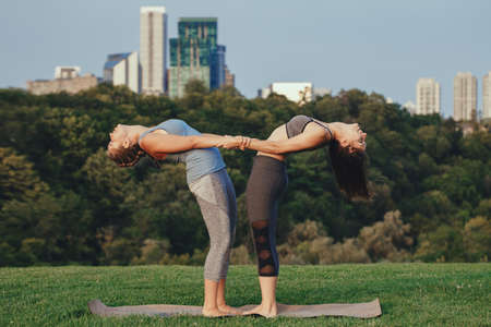 Two young Caucasian women yogi doing balance back stretch acro yoga pose. Women doing stretching workout in park outdoors at sunset. Healthy lifestyle modern activityの写真素材