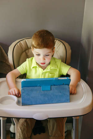 Portrait of cute adorable white Caucasian toddler boy sitting in high feeding chair playing watching...