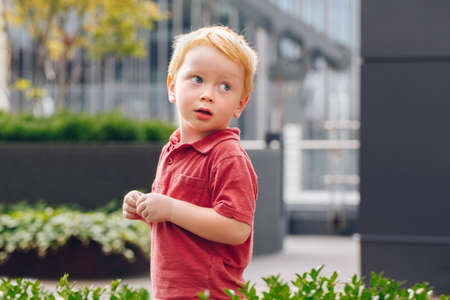 Closeup portrait of cute adorable little red-haired Caucasian boy child in red t-shirt standing in...