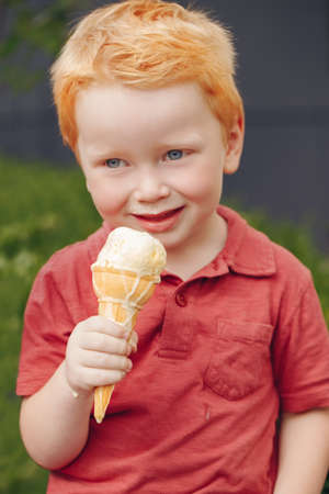 Portrait of cute white Caucasian redhead blond preschool boy with blue eyes in red t-shirt eating licking ice cream in waffle coneの写真素材