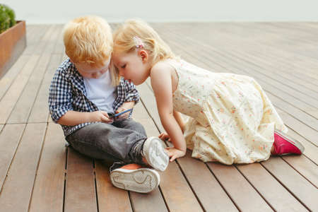 Two cute adorable white Caucasian toddlers boy and girl sitting together and playing games on cell mobile phone digital tablet. Candid lifestyle early development. New technology generation.の写真素材