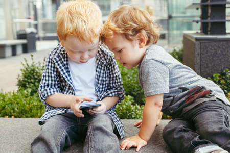 Two cute adorable white Caucasian toddlers boys sitting together and playing games on cell mobile phone digital tablet. Candid lifestyle and early development concept. New technology generation.の写真素材